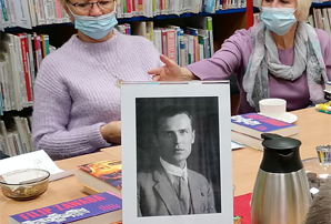 dwie kobiety siedzą przy stole, przed nimi czarno-białe zdjęcie w ramce, w tle książki na regałach