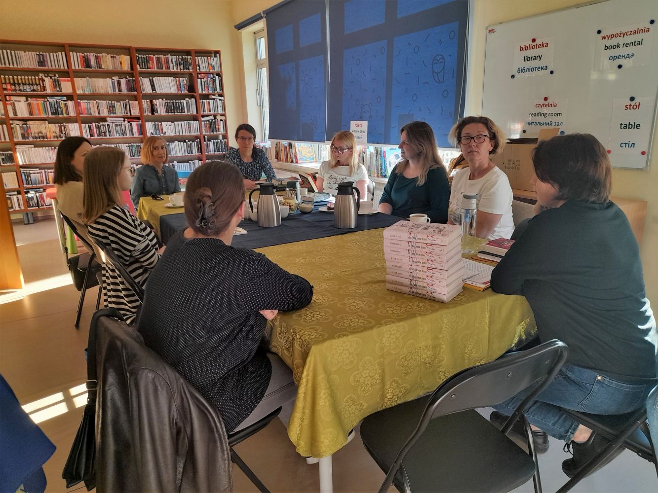 9 kobiet siedzi wokół stołu, na którym są książki, herbata i ciastka. W tle regały z książkami bibliotecznymi