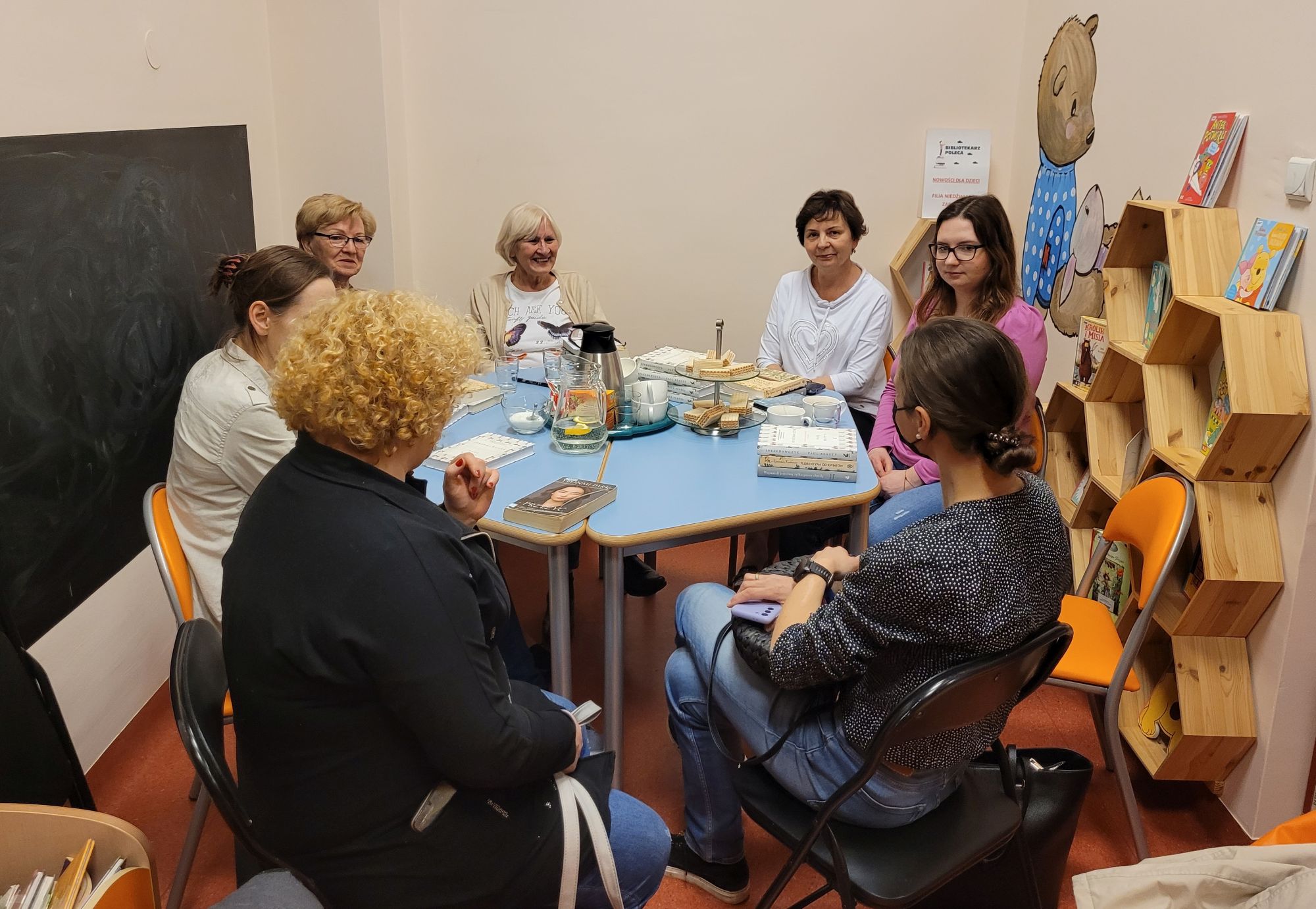 Dyskusyjny Klub Książki przy stole siedzi siedem kobiet, na stole leżą książki, na stole stoi też patera z ciastkami, woda i herbata, w tle widać półkę z książkami