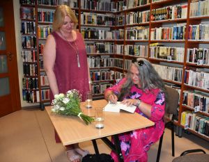 jedna kobieta stoi przy stoliku, przy którym siedzi druga kobieta podpisująca książkę, na stoliku stoją dwie szklanki z wodą i leży bukiet białych kwiatów
