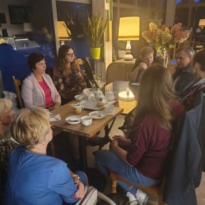 osiem osób siedzi przy stole, na stole stoją filiżanki i talerzyki z ciastem, dookoła kwiaty w doniczkach