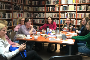 grupka kobiet siedzi przy stole przy ksiązkach i herbacie. W tle biblioteczne regały z książkami