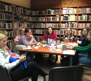 grupka kobiet siedzi przy stole przy ksiązkach i herbacie. W tle biblioteczne regały z książkami