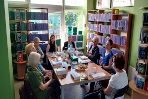 Osiem osób siedzących przy stole. Na stole książki, kubki, ciastka, butelki z wodą, termos, talerzyki. Na półkach w koło stołu stoją książki.