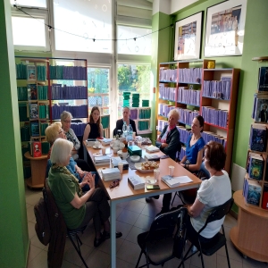 Osiem osób siedzących przy stole. Na stole książki, kubki, ciastka, butelki z wodą, termos, talerzyki. Na półkach w koło stołu stoją książki.