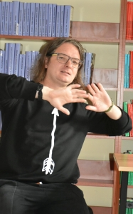 mężczyzna gestykuluje, w tle półki z książkami