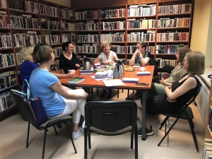 Siedem osób siedzących w koło stołu. Na stole leżą książki, talerzyki i i telefon, stoją kubki, termosy. Przy ścianach w pomieszczeniu stoją wysokie półki z książkami.