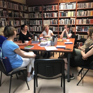 Siedem osób siedzących w koło stołu. Na stole leżą książki, talerzyki i i telefon, stoją kubki, termosy. Przy ścianach w pomieszczeniu stoją wysokie półki z książkami.