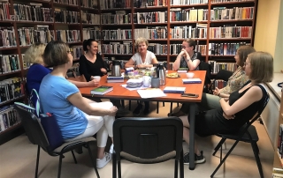 Siedem osób siedzących w koło stołu. Na stole leżą książki, talerzyki i i telefon, stoją kubki, termosy. Przy ścianach w pomieszczeniu stoją wysokie półki z książkami.