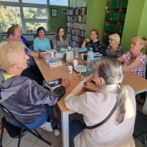 Osiem osób siedzących przy stole. Na stole książki, kubki, ciastka, butelka z wodą, dzbanek, talerzyki. Na półkach w koło stołu stoją książki.