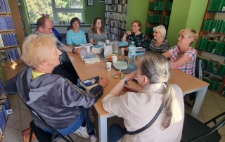 Osiem osób siedzących przy stole. Na stole książki, kubki, ciastka, butelka z wodą, dzbanek, talerzyki. Na półkach w koło stołu stoją książki.