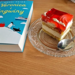 Leżąca na stole książka pod tytułem "Veronica i pingwiny" autorstwa Hazel Piror . Obok talerzyk, na którym jest kawałek ciasta z galaretką i łyżeczka.