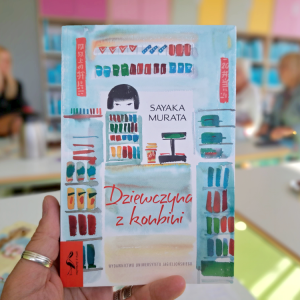 Na pierwszym planie książka trzymana w dłoni. W rozmytym tle widać trzy osoby.