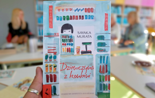 Na pierwszym planie książka trzymana w dłoni. W rozmytym tle widać trzy osoby.