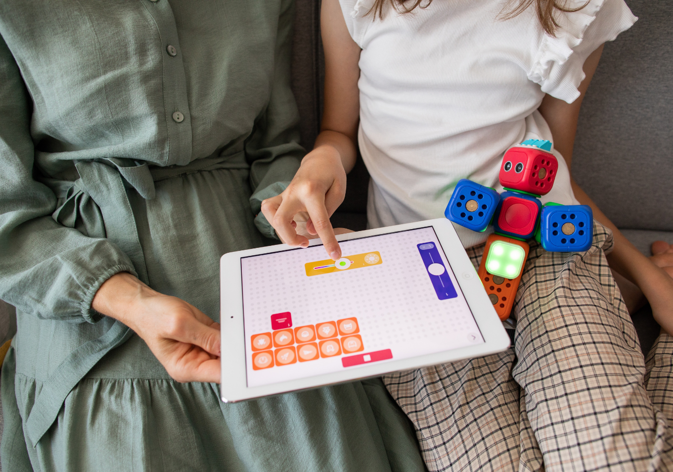 kobieta w zielonej sukience razem z dzieckiem kodują na tablecie, obok dziecka leży czerwono-niebieski robot-zabawka