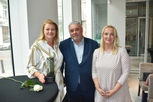 Trzy osoby stoją obok siebie przy wysokim stole. Na stole leży biała róża i stoi statuetka.