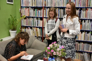 Trzy kobiety. Dwie stoją. Trzecia siedzi przy stoliku i podpisuje otwartą książkę.