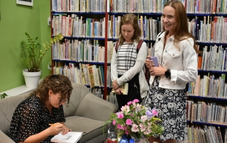 Trzy kobiety. Dwie stoją. Trzecia siedzi przy stoliku i podpisuje otwartą książkę.