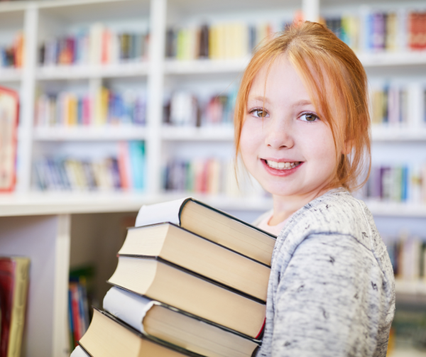 uśmiechnięta dziewczynka trzymająca stos książek, w tle regały z książkami