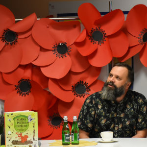 mężczyzna siedzi przy stole na którym jest książka, kawa i woda. W tle czerwone maki