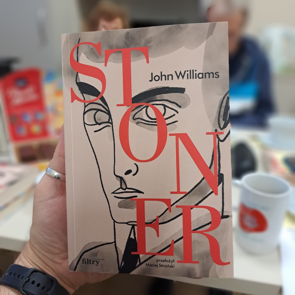 Książka trzymana w dłoni. Na okładce rysunek twarzy mężczyzny. Tytuł Stoner napisany dużymi czerwonymi literami.