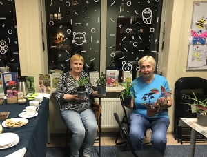 Dwie kobiety siedzą między stołami. Przed nimi stoi czarne krzesło. Kobiety trzymają w dłoniach doniczki z kwiatami. Na jednym ze stołów stoi zastawa. Widać słodycze. Na parapecie za kobietami stoją książki. Na oknach widać naklejki różnych kształtów.