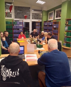 Grupa osób siedzi przy stole. Na stole widać książki, filiżanki, czajniki i ciastka. Na drugim planie regały z książkami.