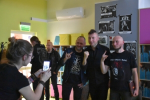 Trzech mężczyzn pozuje do zdjęcia, które telefonem robi kobieta. Za nimi widać plakaty przywieszone na ścianie oraz regały z książkami. W tle stoją dwaj inni mężczyźni.