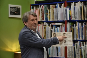 Mężczyzna trzyma w jednej dłoni zdjęcia z opisami. Drugą ręką wskazuje na jeden z opisów. W tle regały z książkami. Na ścianie ramka z tekstem. Za mężczyzną lampka, która rzuca światło na ścianę.