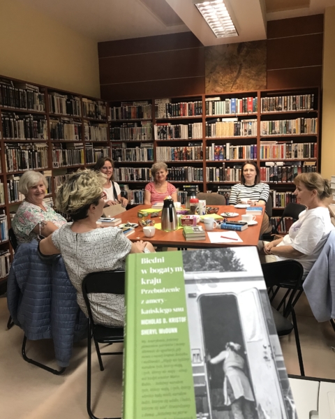 kobiety siedzą dookoła stołu, z tyłu widać regały z książkami. Na pierwszym planie książka.