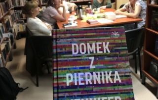 ręka trzyma książkę pod tytułem Domek z piernika. W tle widać kilka osób siedzących wokół stołu w bibliotece