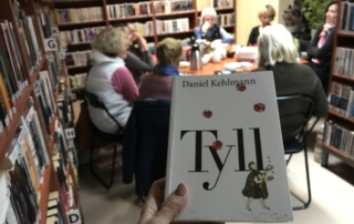 na pierwszym planie ręka trzymająca książkę, w tle kilka osób siedzi przy stole w bibliotece