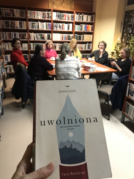 Uwolniona-na stronę kobiety siedzą wokół stołu, za ich plecami widać regały z książkami. na pierwszym planie pokazana książka pod tytułem "Uwolniona"