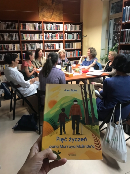 Dłoń z książką, na tle grupa ludzi przy stole.