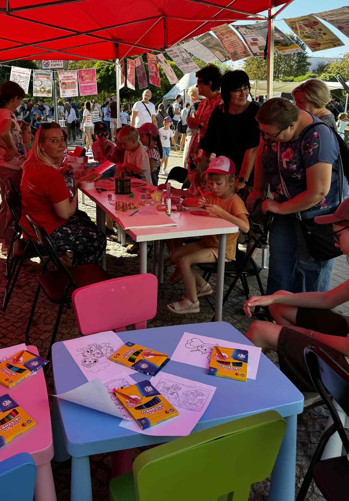 0727485e-3e4b-441c-81ef-50260e5fe205 Przy kolorowych stolikach rozłożone są kredki i kolorowanki, a w tle pod czerwonym namiotem siedzi grupa dzieci