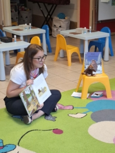 kobieta siedzi na dywanie z książką w rękach, uśmiecha się do dzieci podczas spotkania w bibliotece