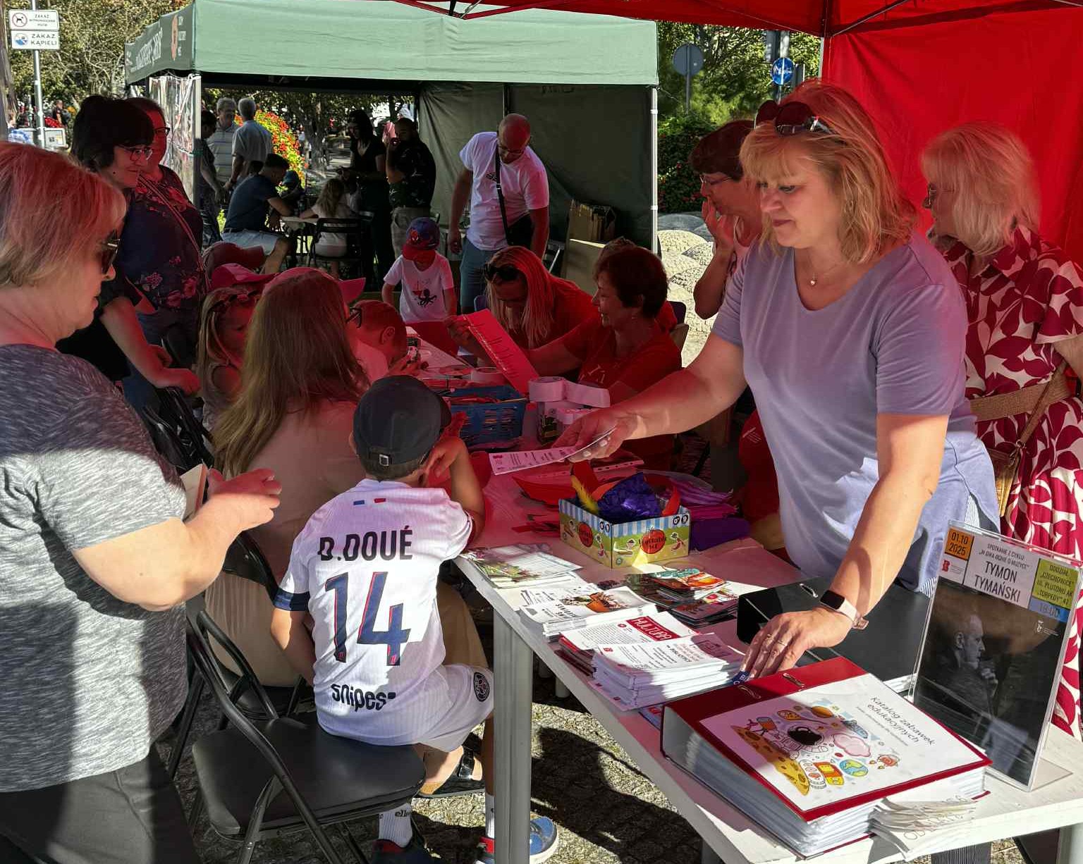 227d07df-b7e6-43f6-aedf-32596e255684 Uśmiechnięta kobieta w fioletowej bluzie wręcza odwiedzającym ulotki i materiały promocyjne podczas pikniku