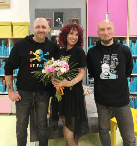 Autorka pozuje z bukietem kwiatów w bibliotece, obok niej stoją dwaj bibliotekarze w ciemnych bluzach