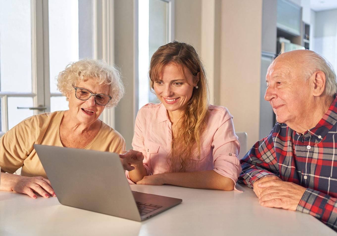 Młoda kobieta siedzi przy stole z seniorką i seniorem, wspólnie oglądają coś na ekranie laptopa.