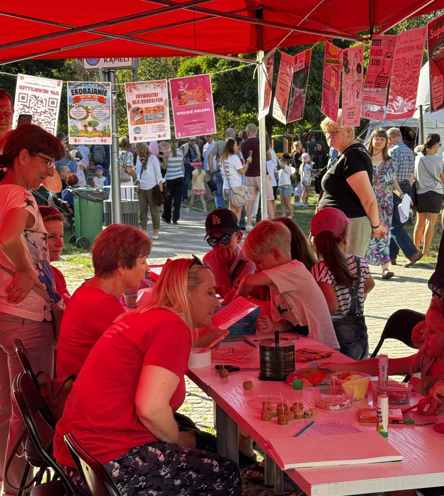 b2f8e528-28cb-4045-b773-0cac23d60da7 Grupa dzieci siedzi przy stolikach pod czerwonym namiotem i zajmuje się kolorowaniem oraz wycinaniem w ramach warsztatów plastycznych.