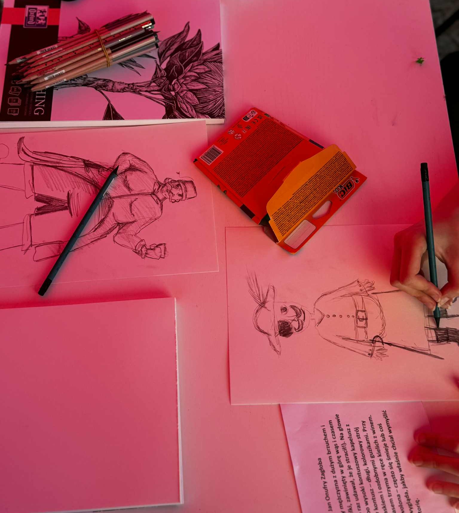 be2776c6-9878-4e71-8ae5-f0bf3647bec0 Na stole leży kilka rysunków karykatur, obok nich rozłożone są ołówki używane do szkicowania