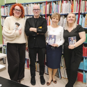 Cztery osoby stoją przed regałami z książkami; trzy kobiety i mężczyzna w czarnej marynarce pozują do wspólnego zdjęcia, trzymając książki