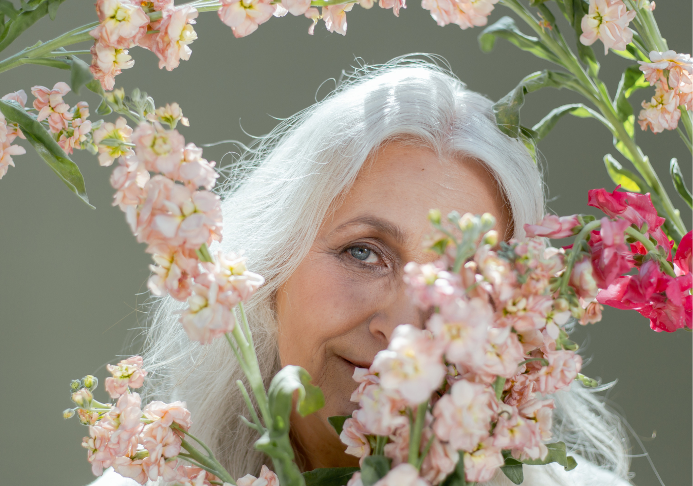 Starsza kobieta o siwych włosach spogląda zza różowych kwiatów, które częściowo zasłaniają jej twarz