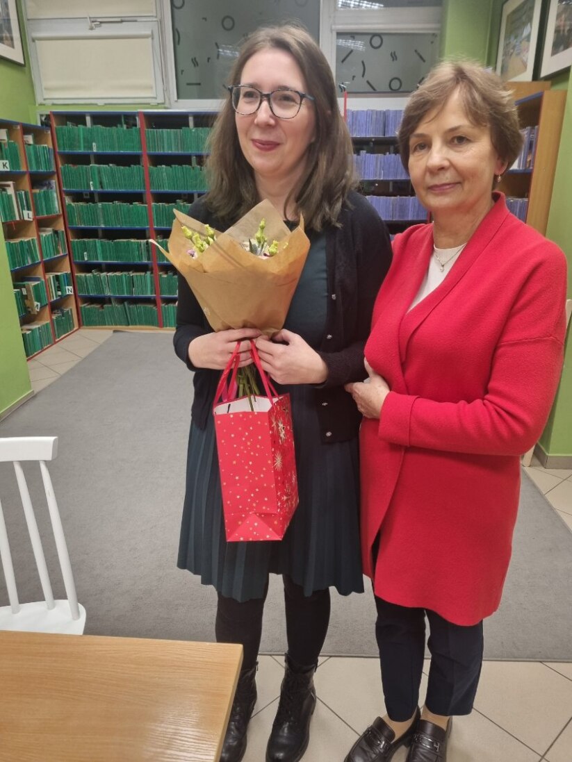 image000000_1764143808449 Dwie kobiety z bukietami kwiatów stoją przed regałem pełnym książek.