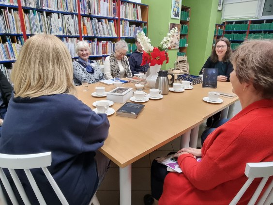 image000000_1764143886614 Grupa ludzi siedzi przy stole w bibliotece, otoczona książkami