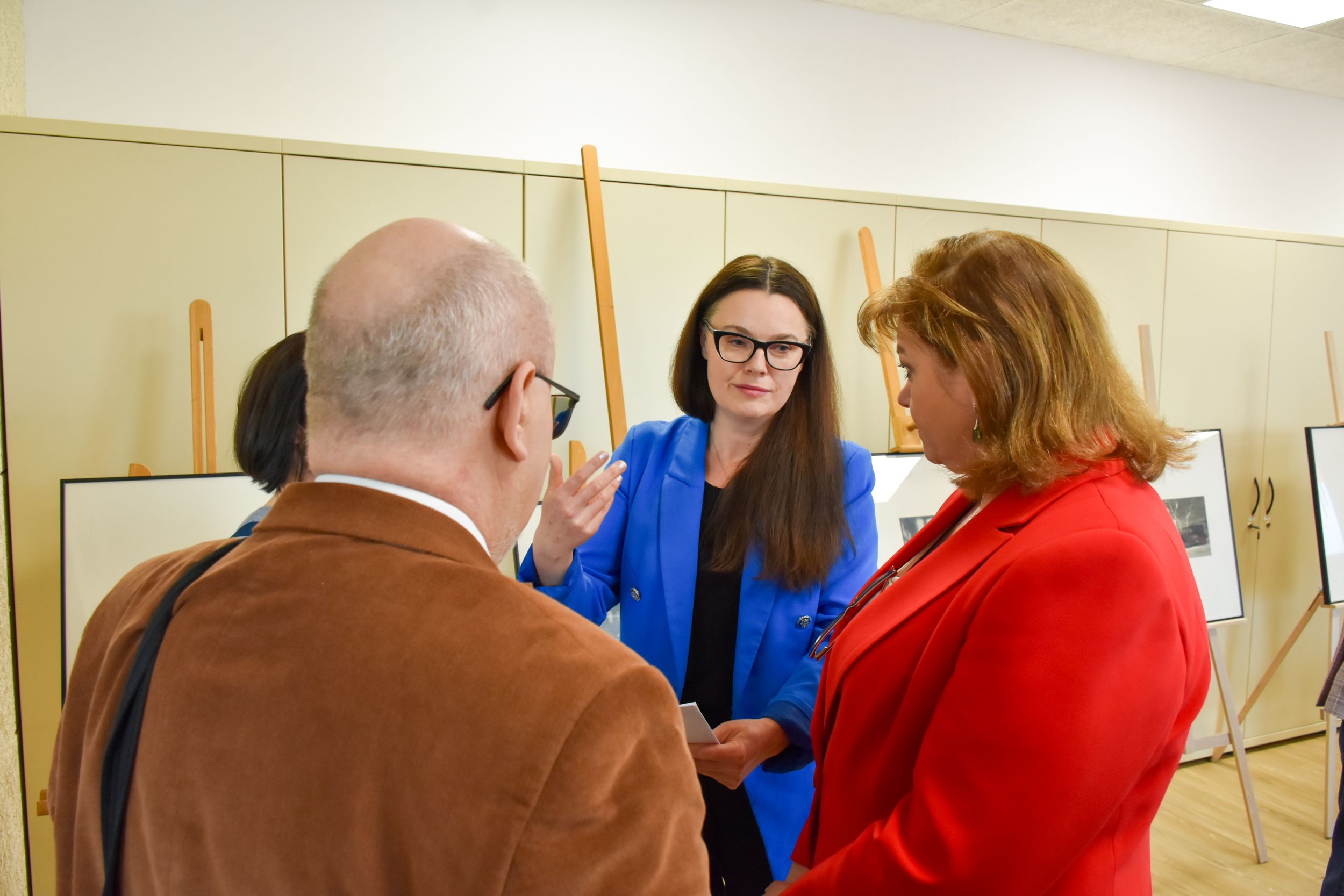 DSC_1924 Grupa czterech osób dyskutuje na temat książek. Na dalszym planie stoją sztalugi ze zdjęciami. Kobieta w niebieskim żakiecie ma uniesioną do góry rękę i rozmawia z kobietą w czerwonym żakiecie.