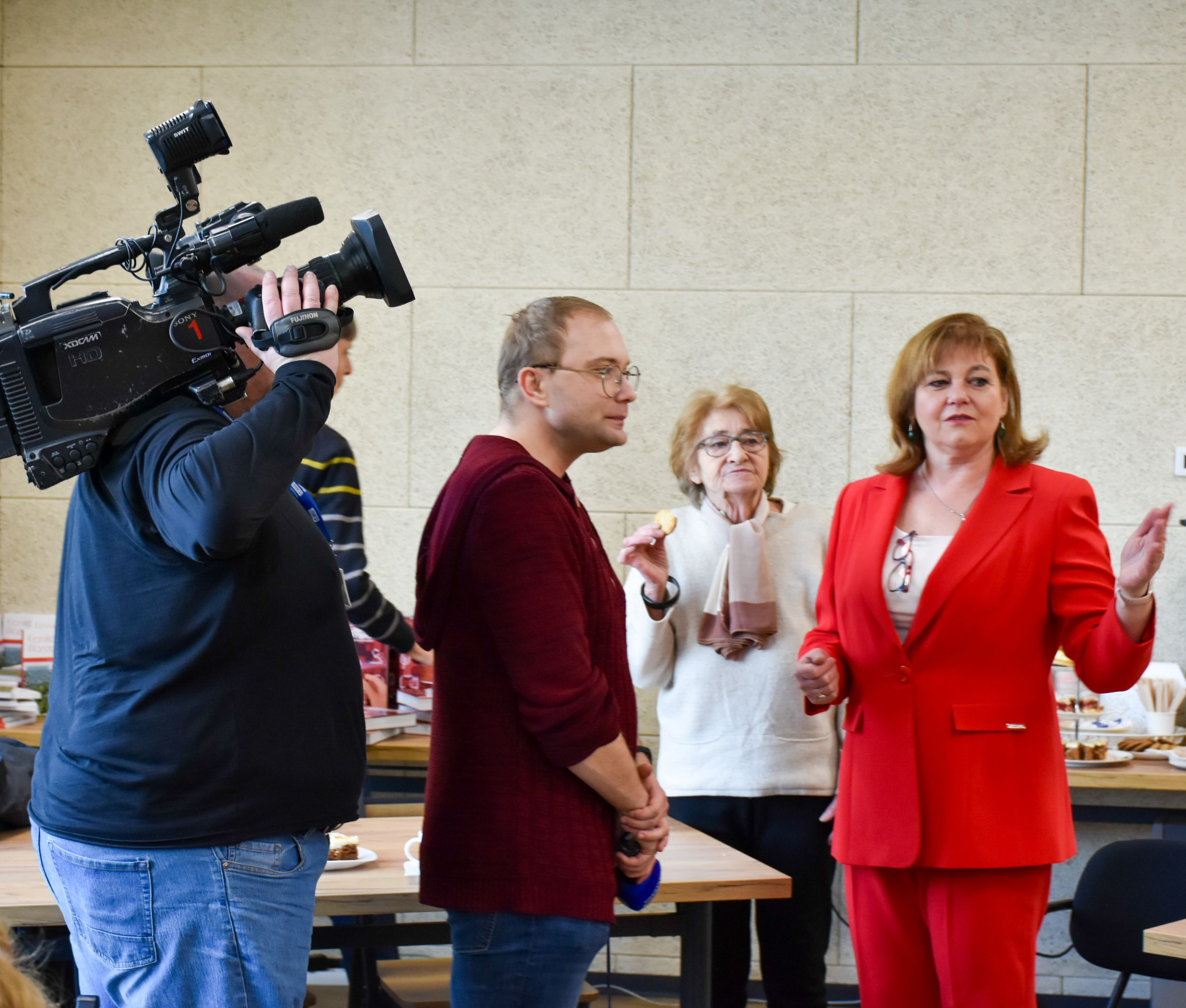 DSC_1940 Po lewej stronie stoi profilem mężczyzna z kamerą na swoim barku następnie mężczyzna w okularach i bordowej bluzie. Po prawej stronie stoi kobieta w białej bluzeczce i okularach trzyma ciastko druga kobieta w czerwonym żakiecie wskazuje dłonią w dal