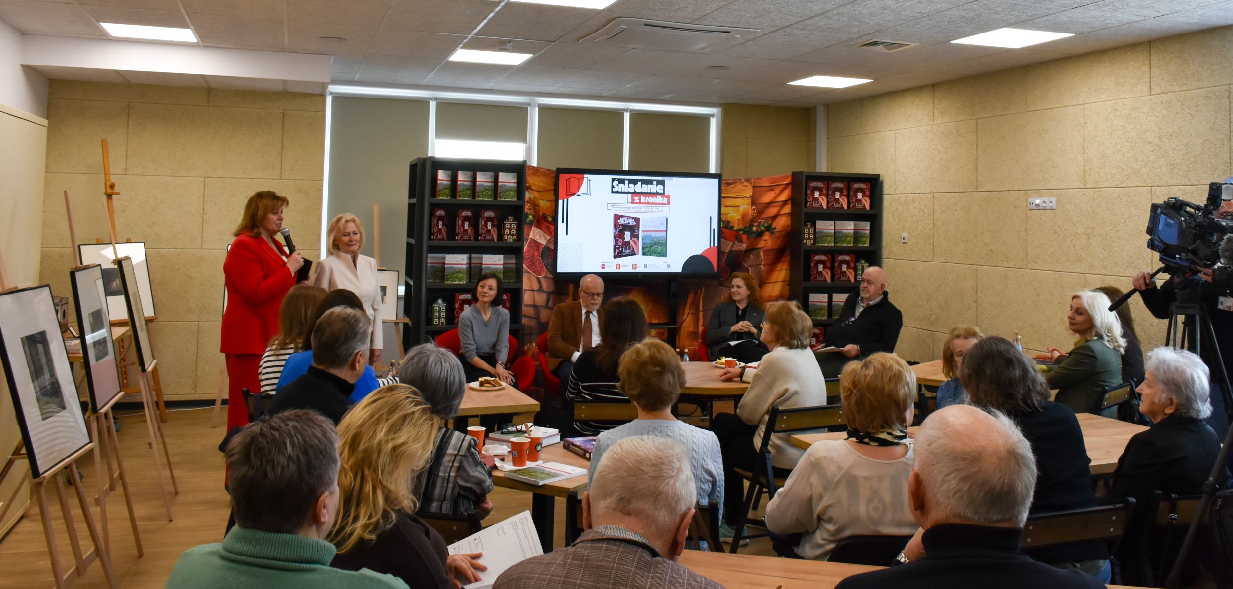 DSC_1972 grupa osób zwróconych tyłem siedzących przy stolikach i słuchających kobiety w czerwonym żakiecie przemawiającej przez mikrofon obok której stoi pani w kremowym kostiumie ze stójką. Na dalszym planie siedzą dwie kobiety i dwóch mężczyzn na tle tablicy oraz dwóch regałów z książkami po obu jej stronach. Z lewej strony stoją sztalugi ze zdjęciami