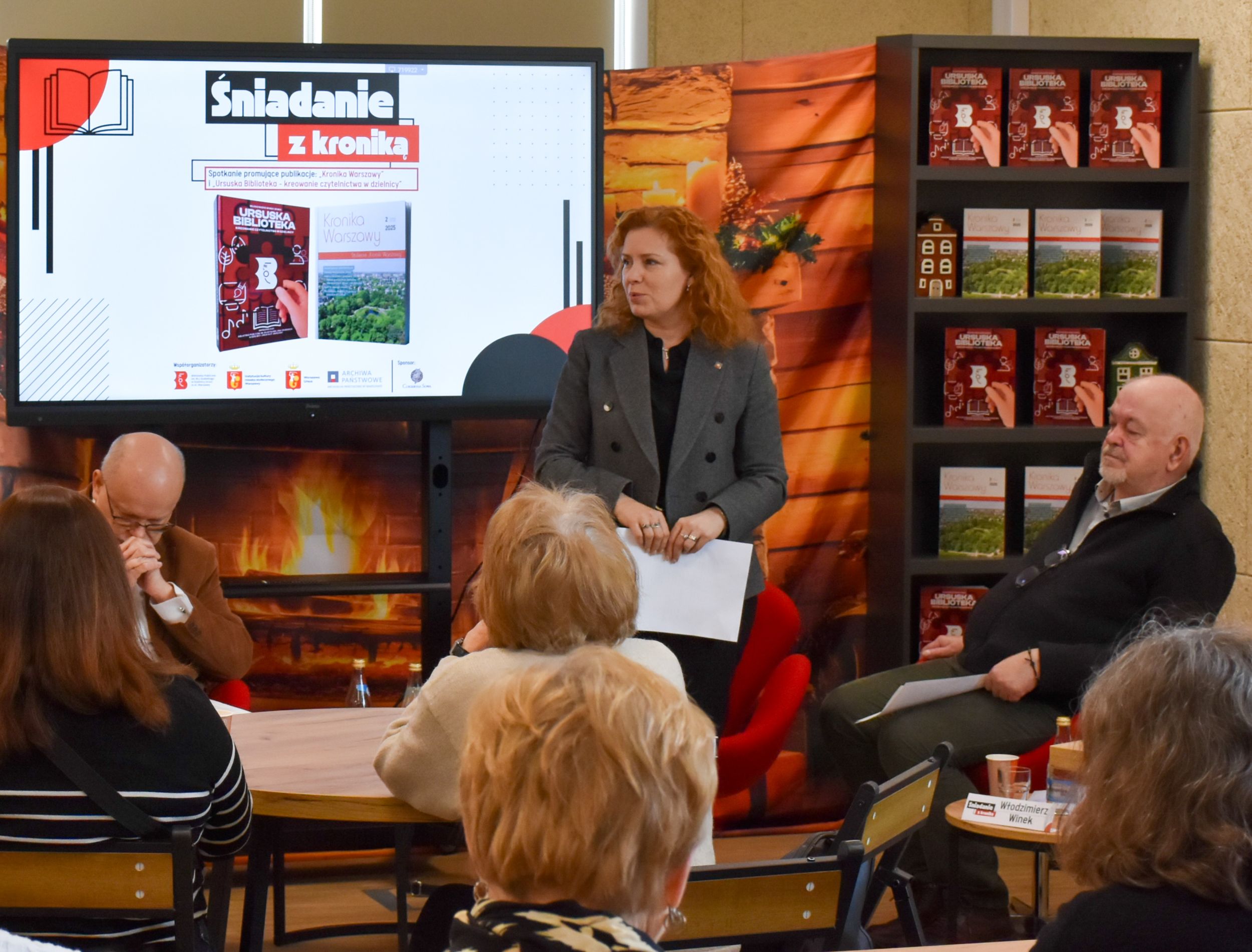 DSC_1983 Kobieta o rudych kręconych włosach w szarym żakiecie z czarnymi guzikami na tle slajdu przedstawiającego Śniadanie z kroniką przemawia do siedzących ludzi