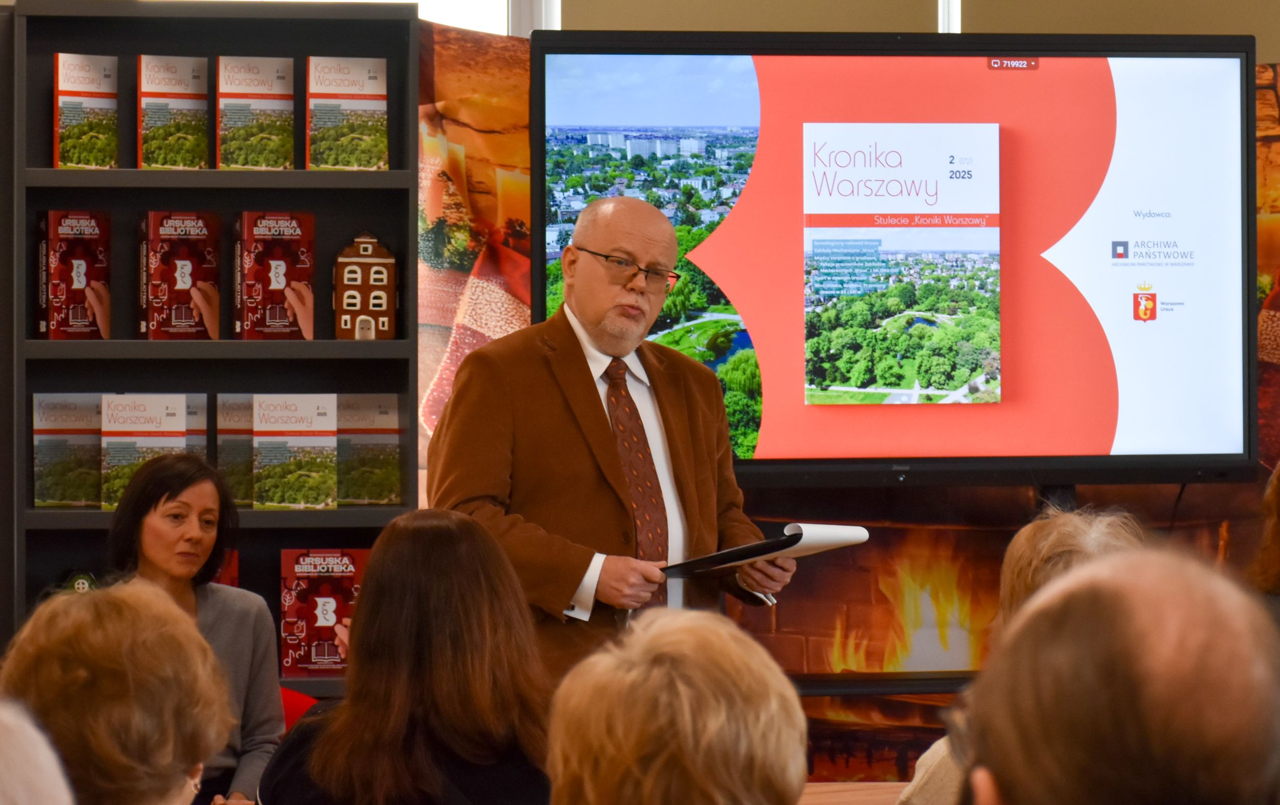 DSC_1999 Na zdjęciu stoi mężczyzna w garniturze białej koszuli krawacie i okularach trzymający notatnik i przemawiający do siedzących ludzi. W tle regał z ursuskimi książkami oraz slajd na tablicy przedstawiający Kronikę Warszawy nr 2 z 2025 roku